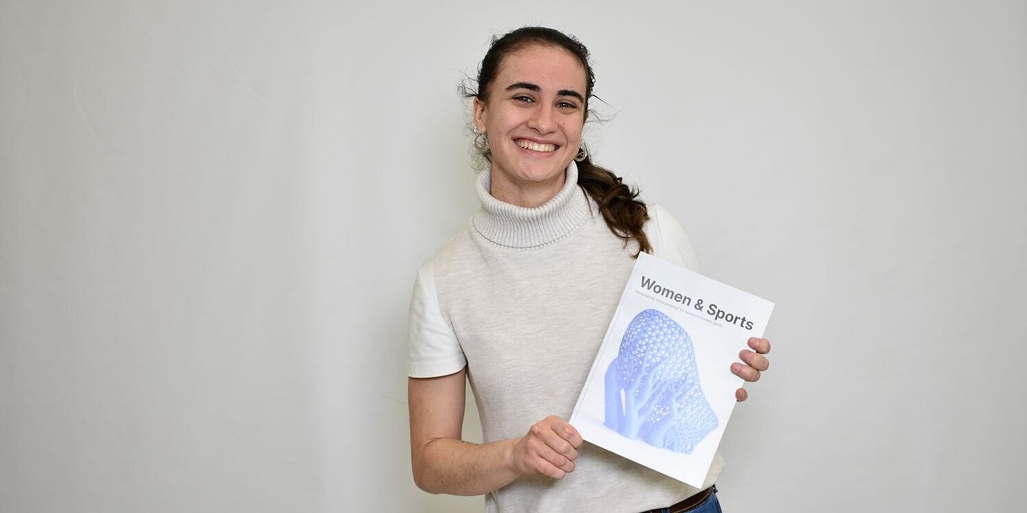 Ein Porträt von Beatriz Jerez Arnau, die die Abschlussarbeit (ihre Bachelorarbeit) in der Hand hält und in die Kamera lacht. 