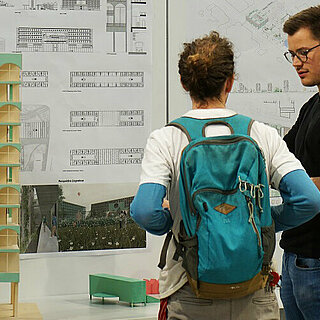 Zwei Personen im Gespräch vor einem Tisch mit Modellen und Stellwänden mit Architekturplänen.
