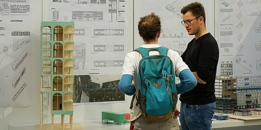 Zwei Personen im Gespräch vor einem Tisch mit Modellen und Stellwänden mit Architekturplänen.
